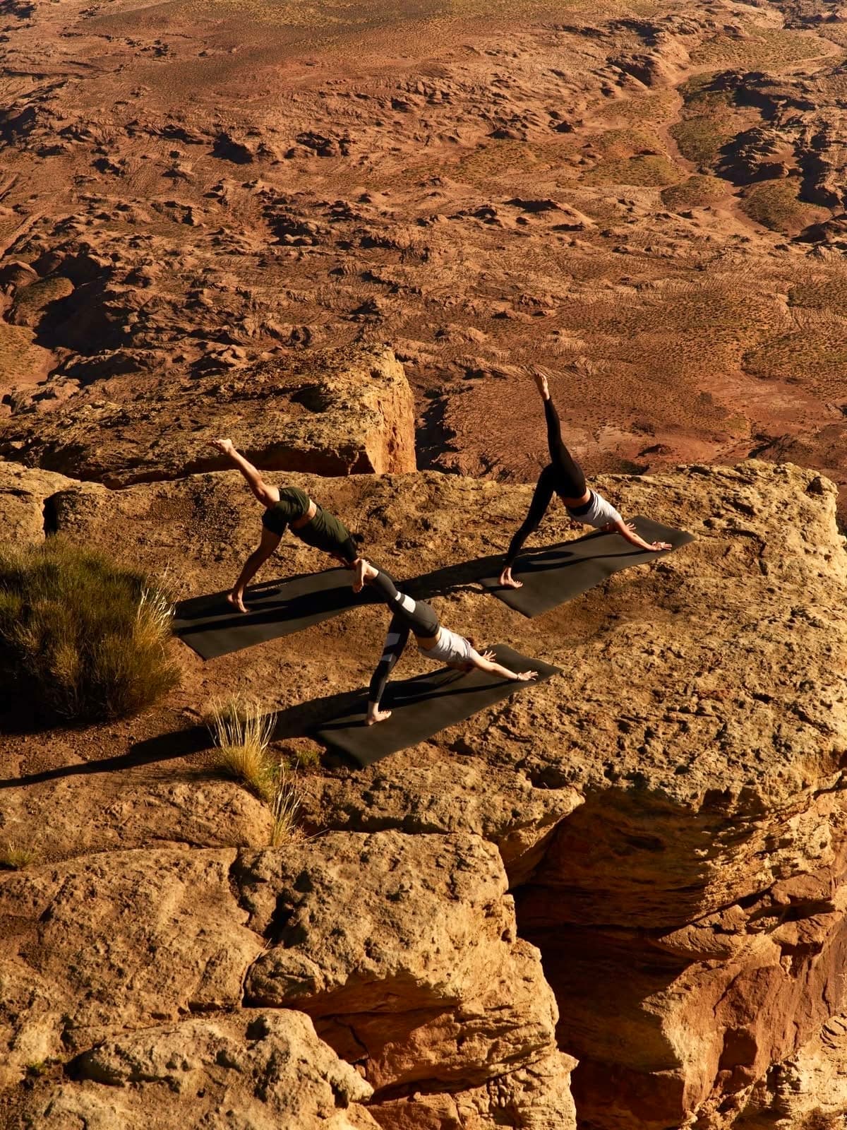 Three people practising yoga on mats along a dramatic red-rock cliff edge