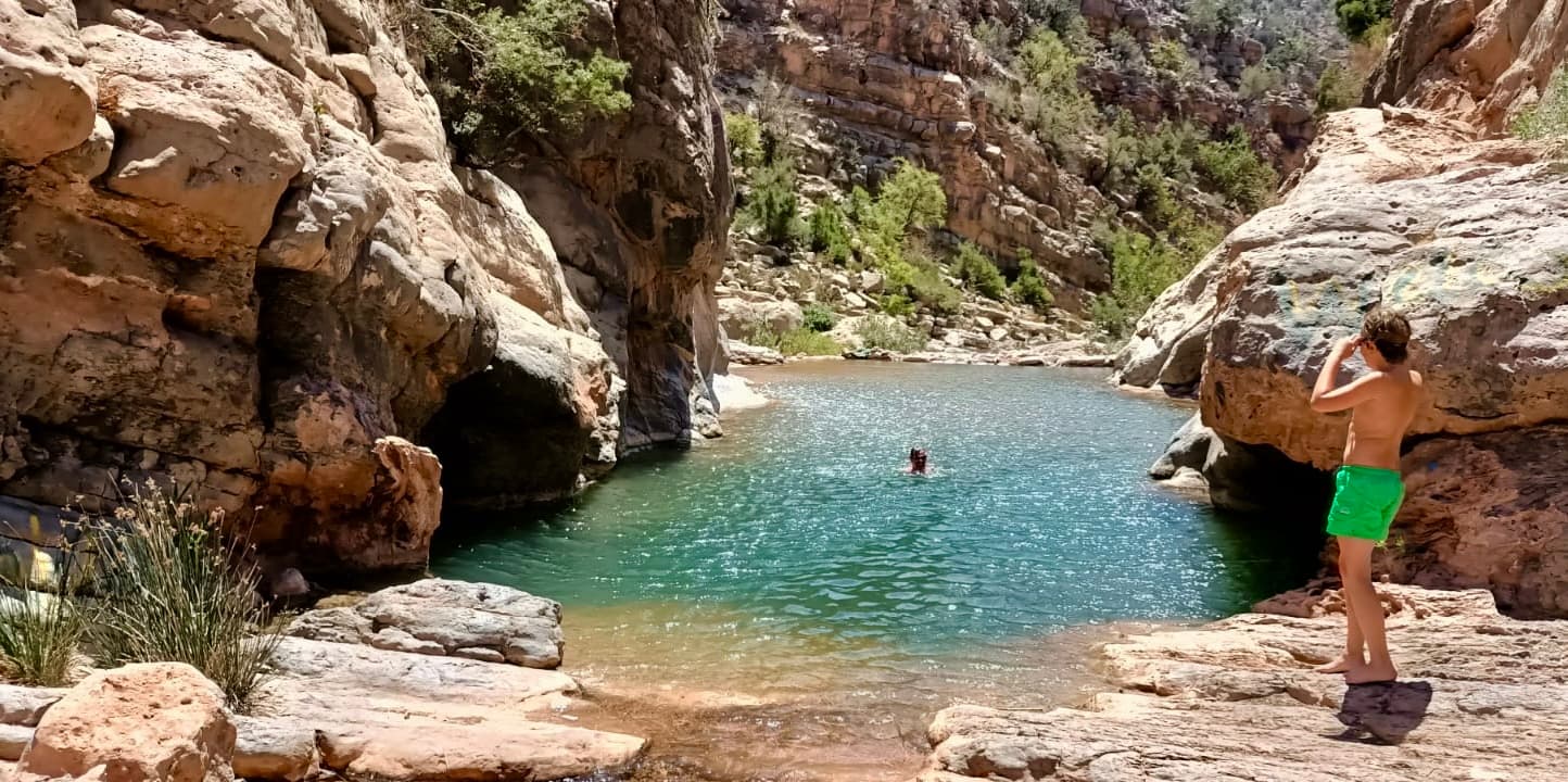 A turquoise natural pool between red rock canyon walls, with two people swimming and standing on the rocks